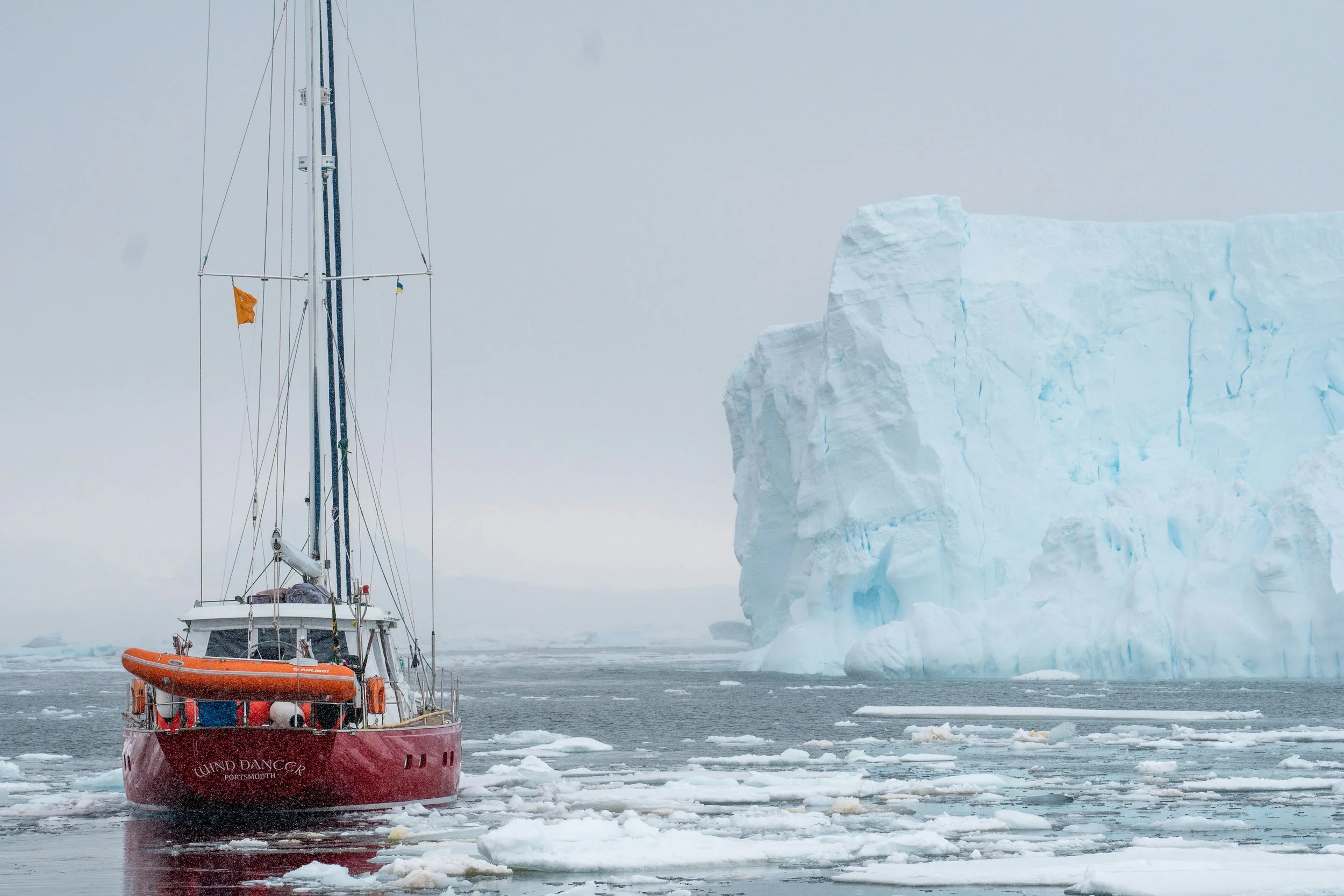 A red sailboat named 'Wind Dancer' sailing among icebergs and ice floes in icy waters, with a large blue iceberg in the background.