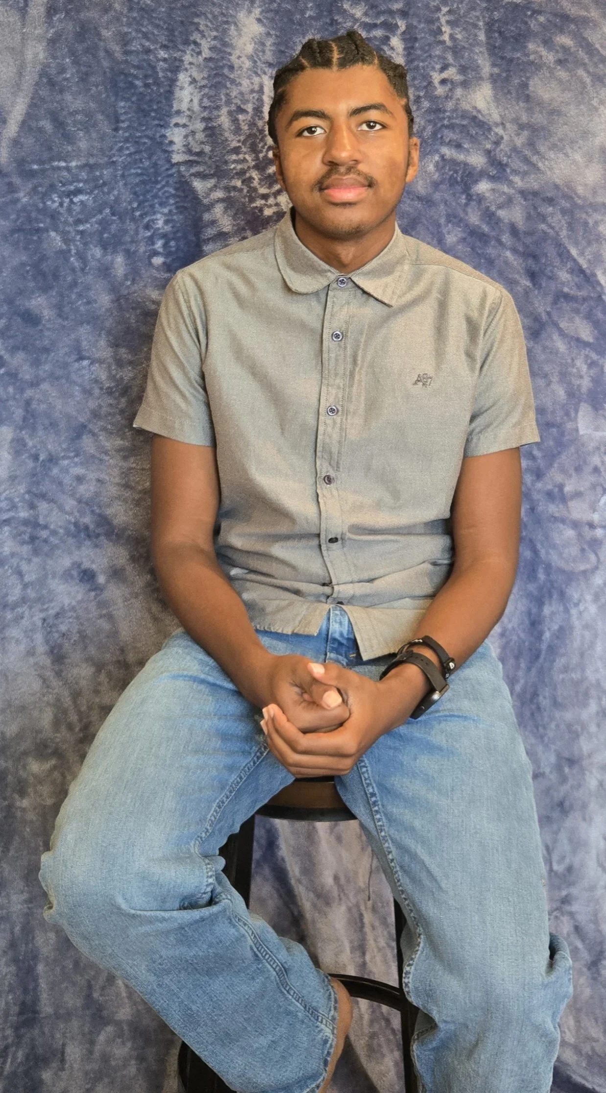 A young man with dark hair styled in cornrows, sitting on a stool against a mottled blue background, wearing a gray short-sleeved button-up shirt and light blue jeans, with his hands clasped in his lap.