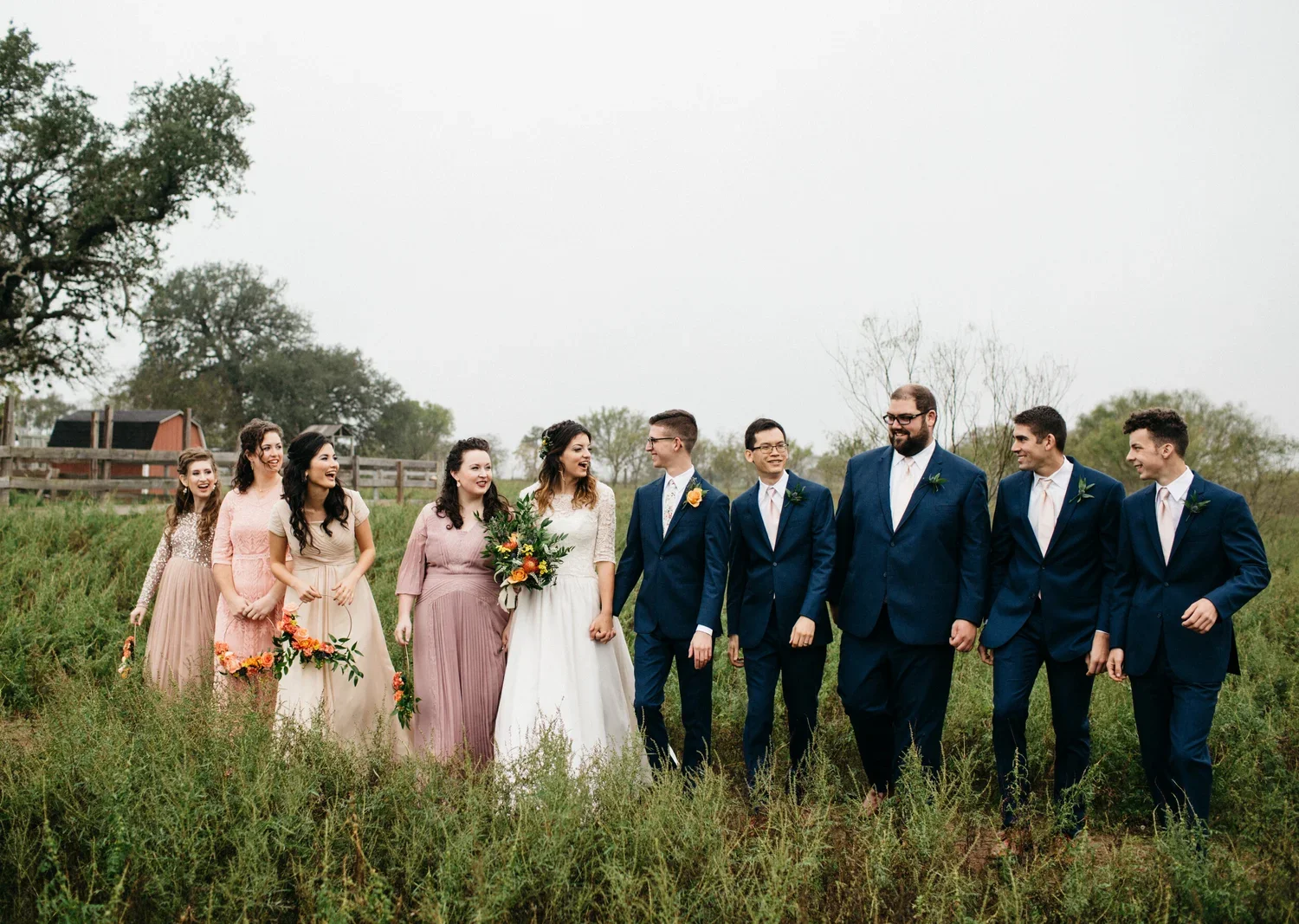 A wedding party outdoors on a cloudy day walking through a grassy field. The group includes women in pastel and white dresses, some holding bouquets, and men in matching blue suits with pink ties and boutonnières. They are smiling and looking at each