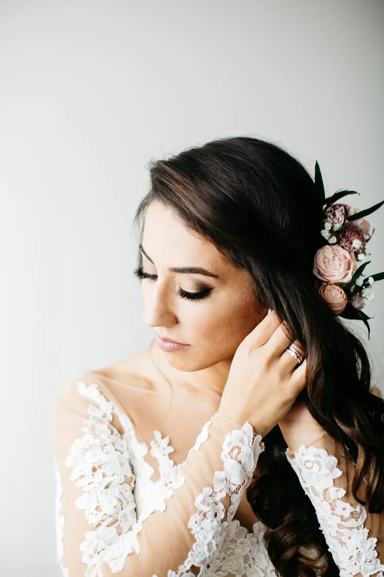 A bride with brunette hair styled in loose curls, wearing a lace wedding dress with floral embroidery, and a floral hairpiece with pink and white roses, adjusting her earring and looking downward.