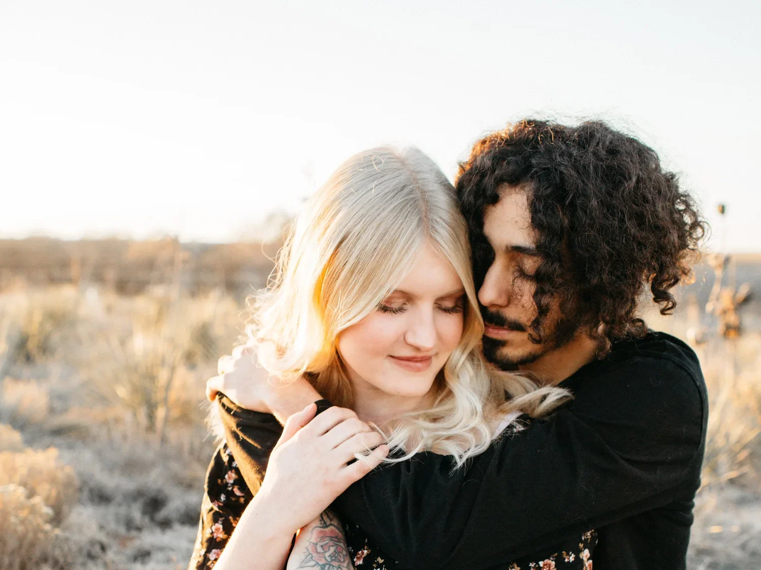 A young couple hugging in a desert landscape at sunset, with their eyes closed.