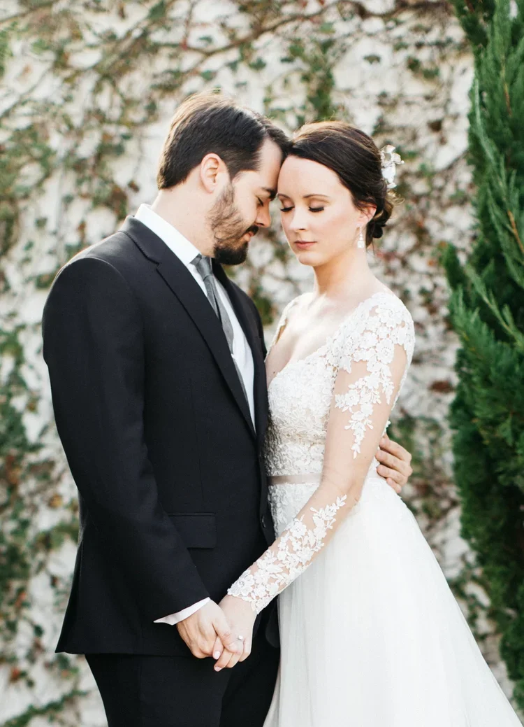 A bride and groom stand closely with their foreheads touching and eyes closed, holding hands outdoors during a wedding. The bride wears a white lace wedding gown with long sleeves and floral lace details, while the groom is dressed in a black suit an