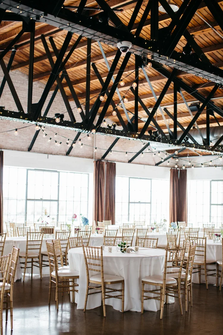 An indoor event space with large windows, round tables with white tablecloths, gold Chiavari chairs, and floral centerpieces. String lights are hung across the exposed wood ceiling with black metal beams.