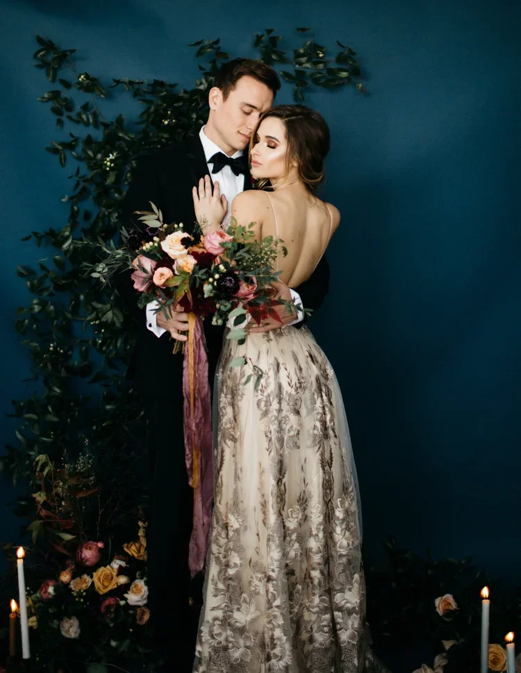 A romantic couple dressed in formal wedding attire embraces each other, with the woman holding a colorful bouquet of flowers. The background features dark green foliage and candles on the floor.