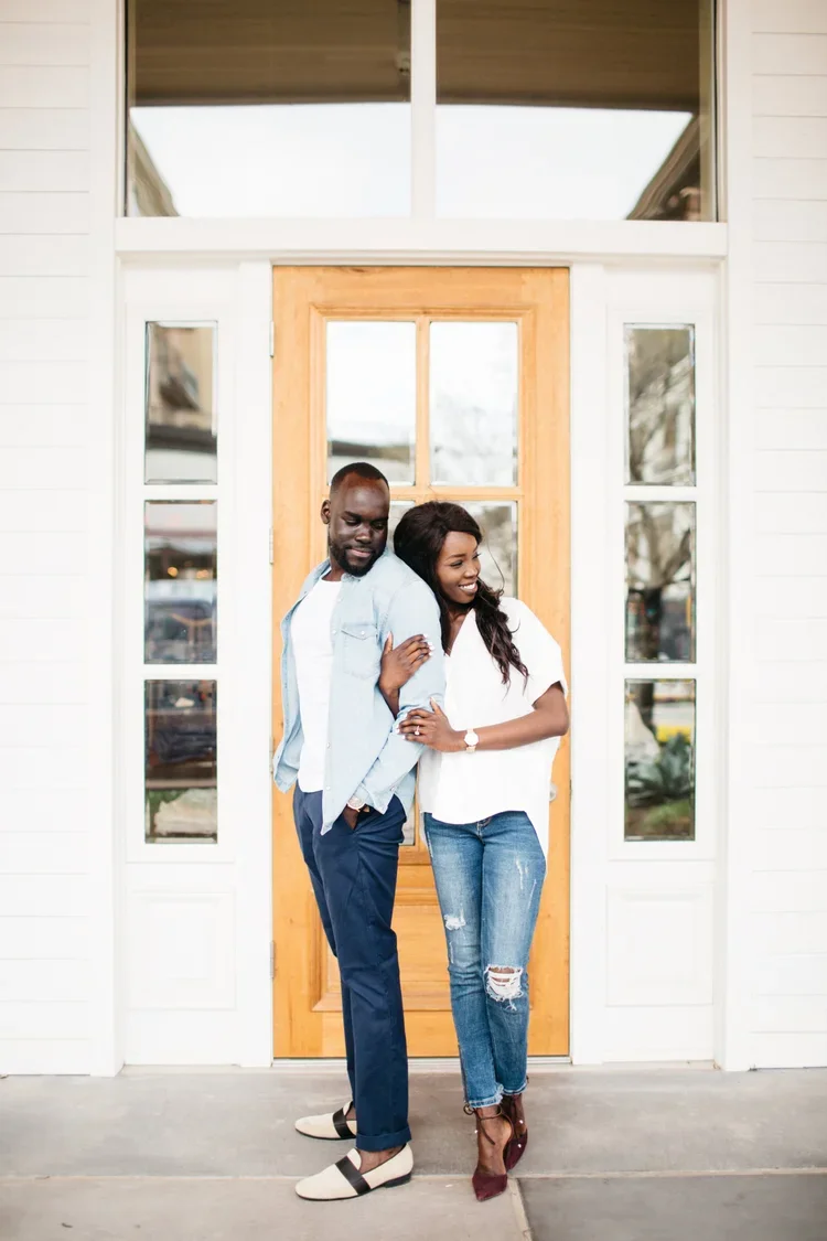 A smiling couple stands in front of a house door, with the woman leaning on the man's shoulder. The man wears a light denim jacket, white T-shirt, and dark pants. The woman wears a white top, ripped jeans, and maroon heels.