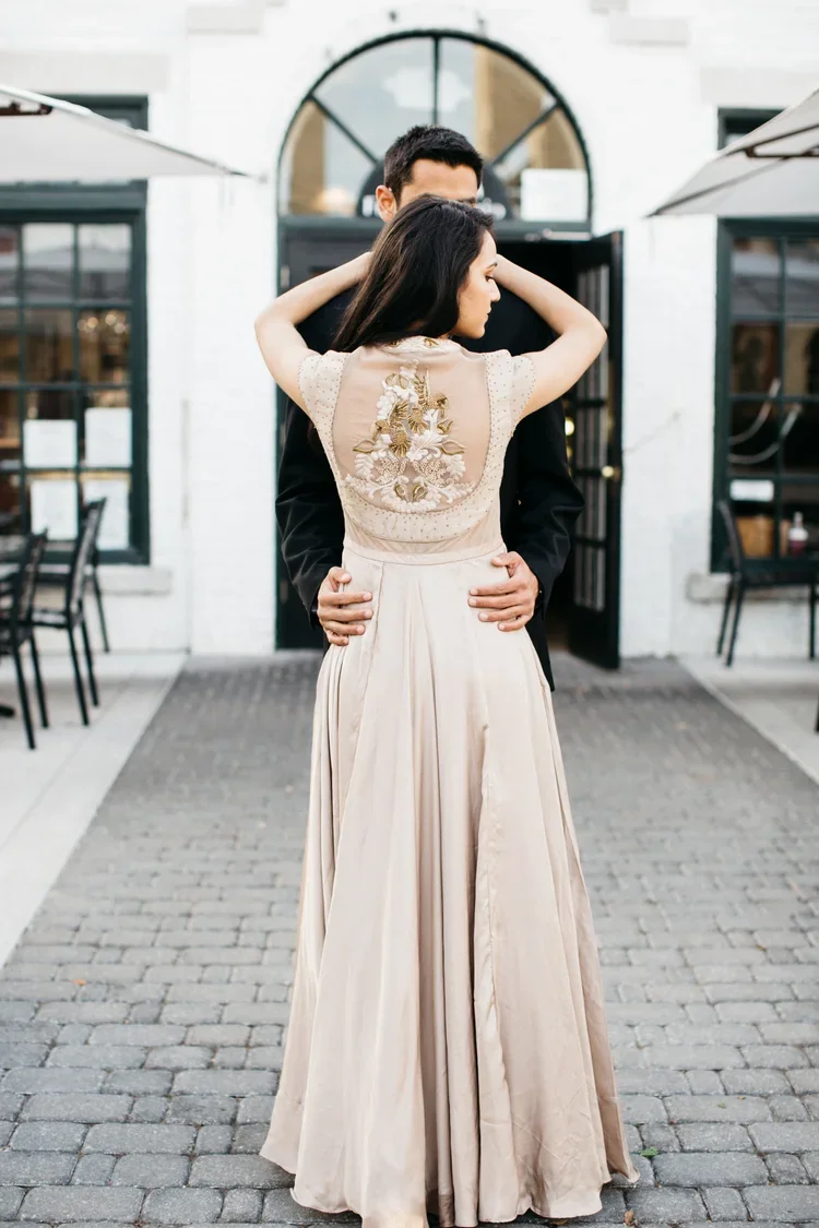 A couple dancing closely outside a building with large windows and an arched door, the woman in a long beige dress with embroidery on the back and the man in a black suit.