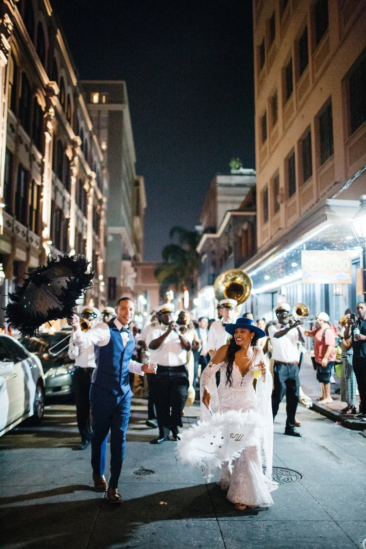 A couple dressed in wedding attire walking and dancing in a city street at night, accompanied by a band playing brass instruments.
