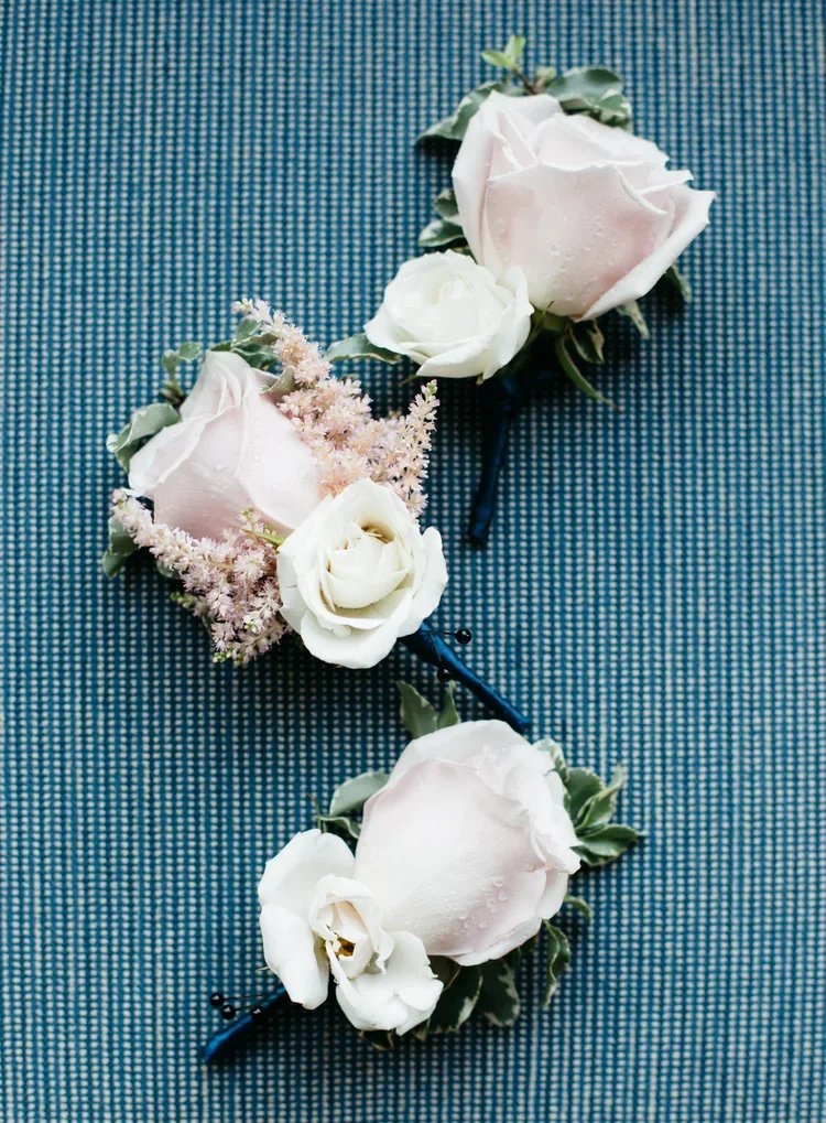 Close-up of a boutonniere with white and pale pink roses, green leaves, and pink filler flowers on a textured blue fabric background.