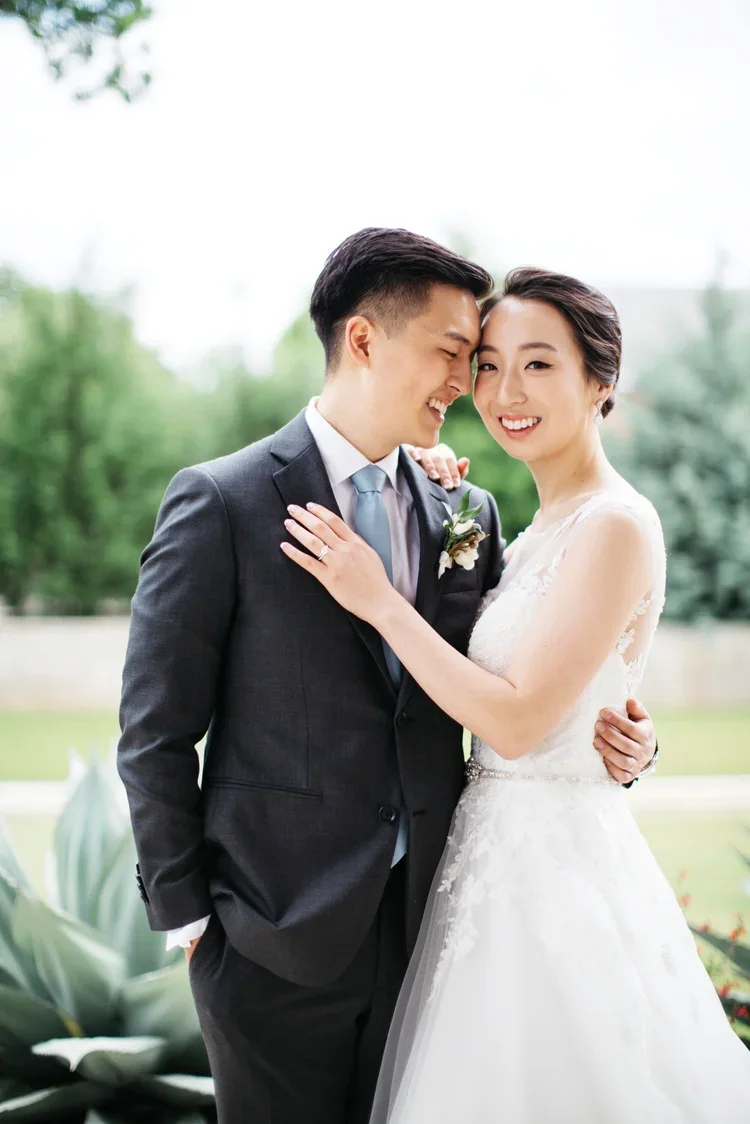 A happy bride and groom embrace outdoors during their wedding, smiling and looking at each other, with trees and greenery in the background.