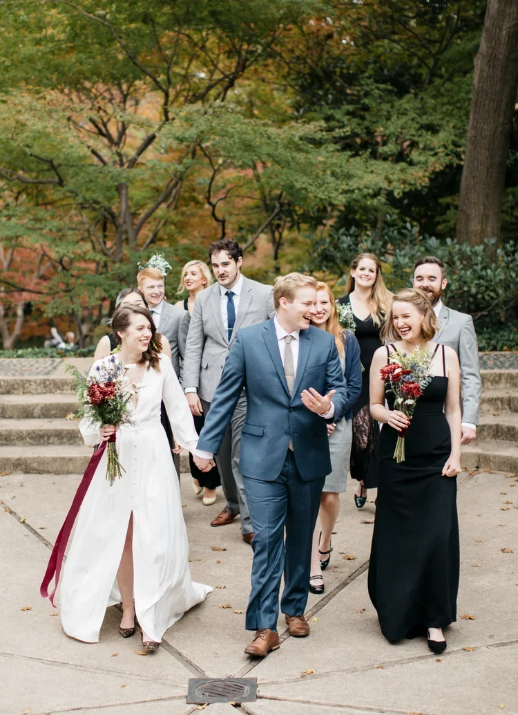 Group of smiling people in formal attire walking outdoors in autumn park, carrying flowers.