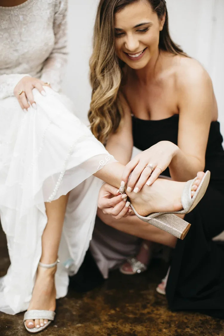 A woman helping another woman put on a high-heeled sandal during a wedding preparation.