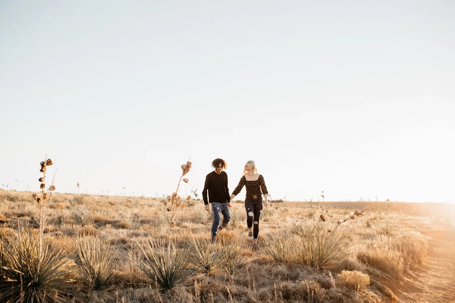 Two women walking hand in hand through a dry, grassy desert landscape at sunset.