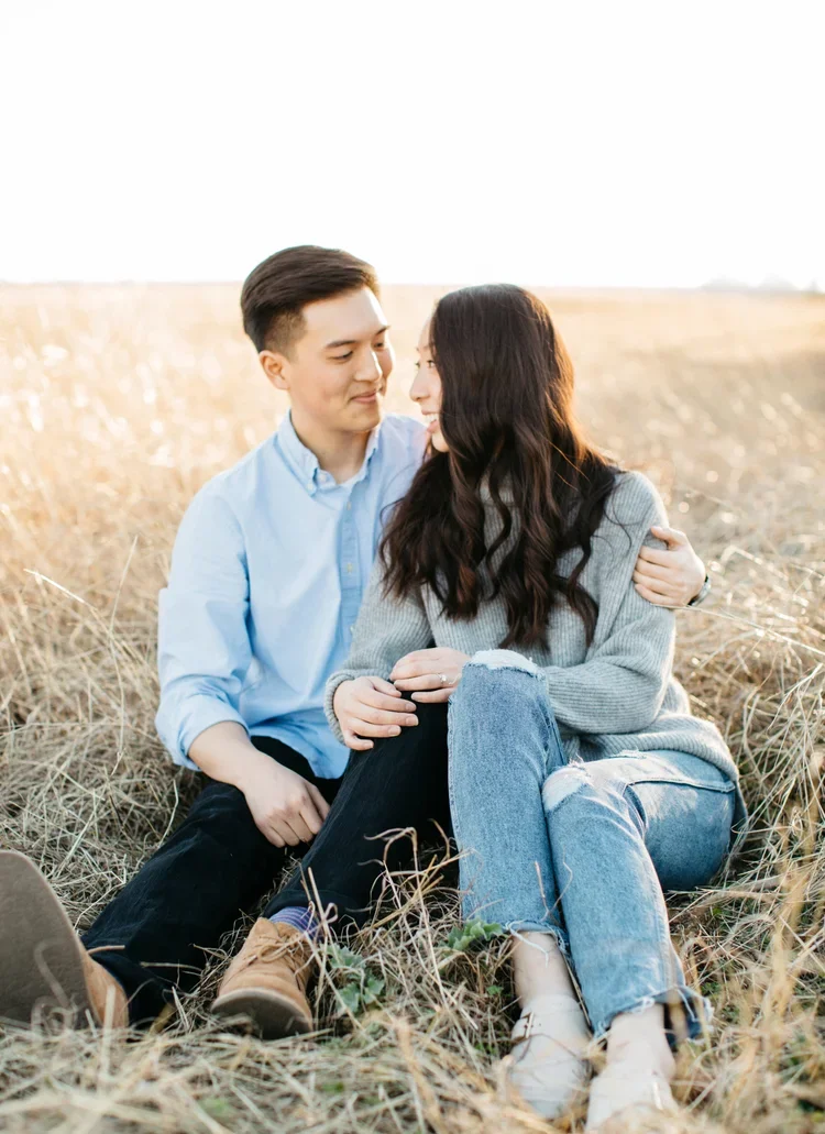 A young couple sitting close together in a field of tall, dry grass, smiling and looking at each other during sunset.