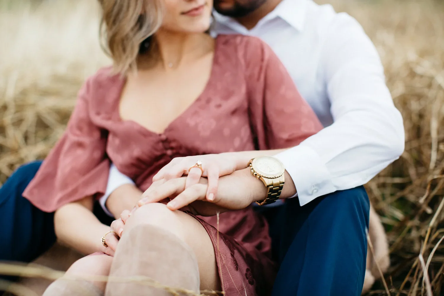 A couple sitting outdoors in a field, with the man holding the woman's hand, showing an engagement ring and wearing a gold watch.