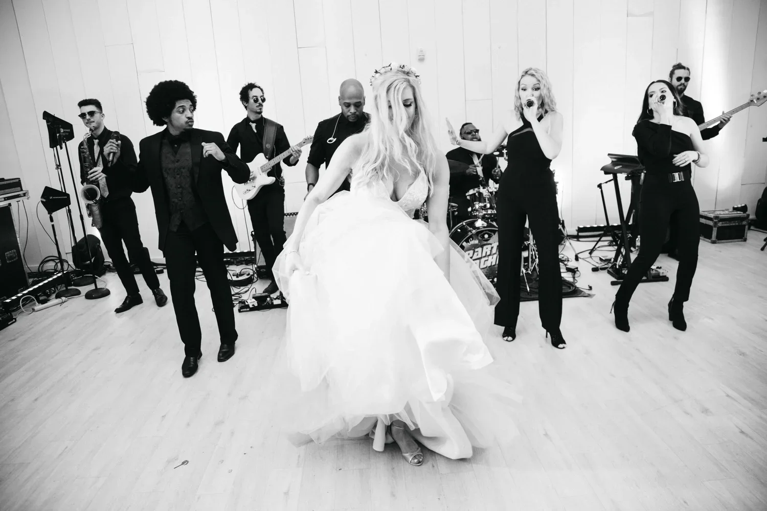 A woman in a wedding dress dancing in front of a band during a celebration, with a band playing instruments behind her in a room with light-colored walls.