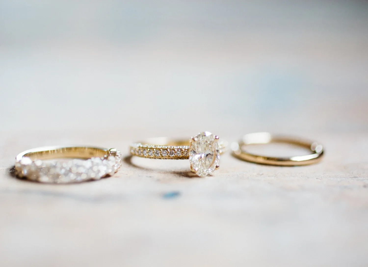 Three gold wedding rings lined up on a surface with a blurred background, one with a large central diamond and two with smaller diamonds.