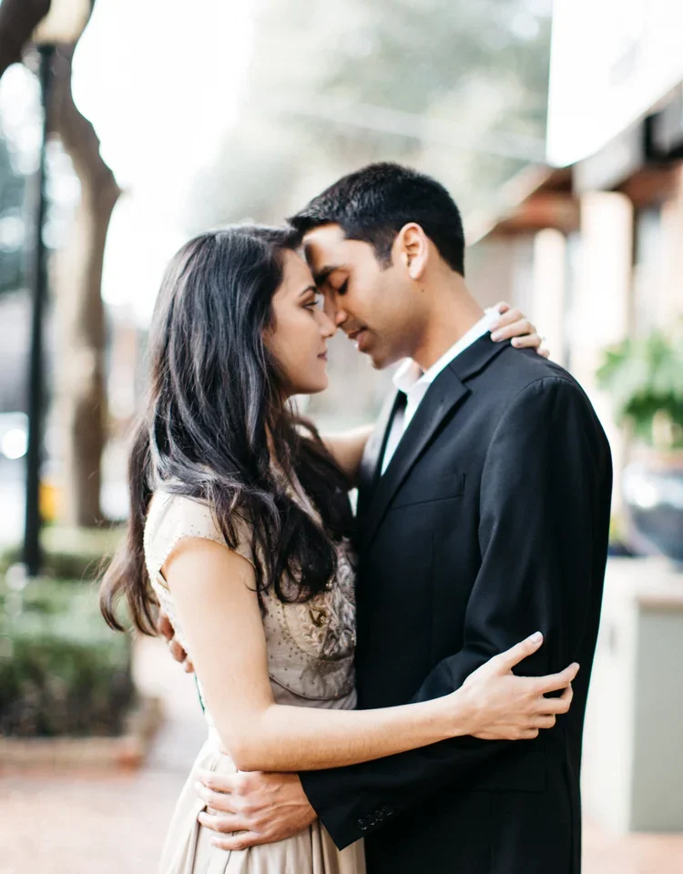 A young couple with light skin embracing outdoors with noses touching, eyes closed, and arms around each other.