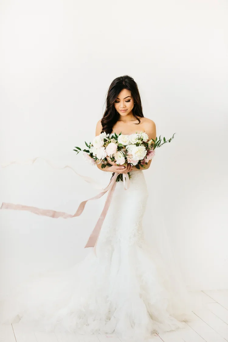 Bride holding a large bouquet of white and pink flowers in front of a plain white background, wearing a strapless white wedding gown.