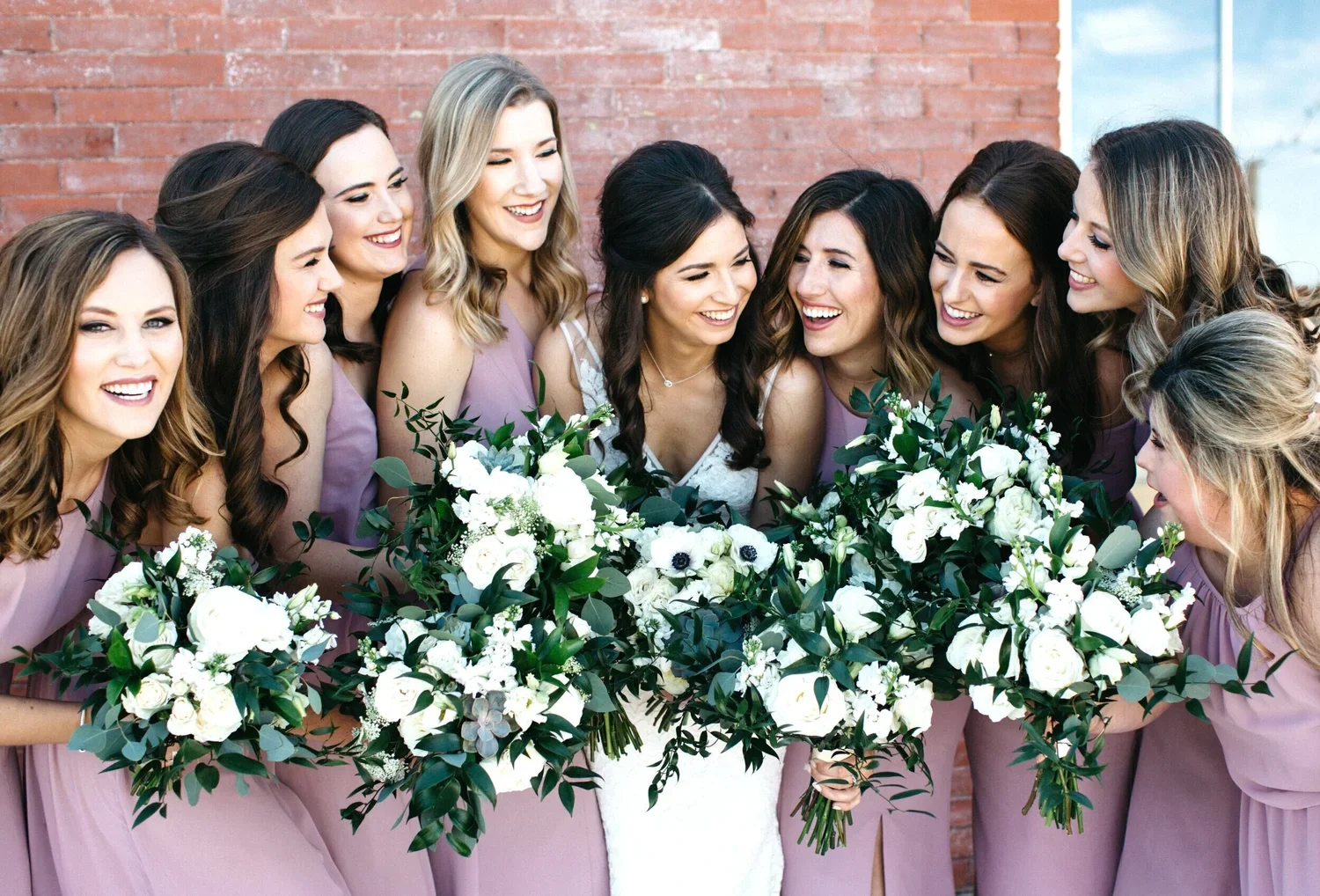 A group of nine women, dressed in light purple dresses, gathered around a bride in her wedding gown, holding large bouquets of white flowers and greenery, laughing and smiling together.