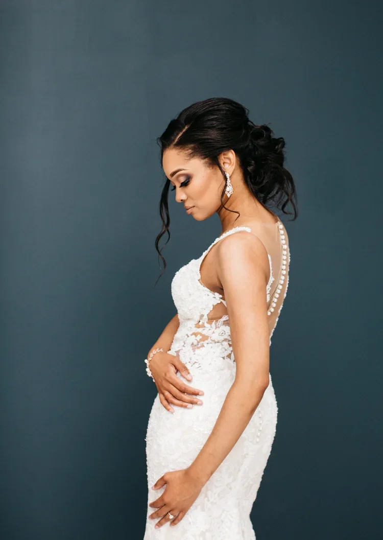 Woman in a white lace wedding dress with pearl details, looking down, against a plain dark background.
