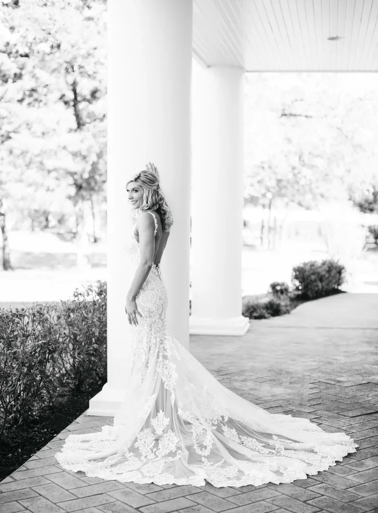Black and white photo of a woman in a wedding dress standing outdoors next to a white column, with trees in the background.