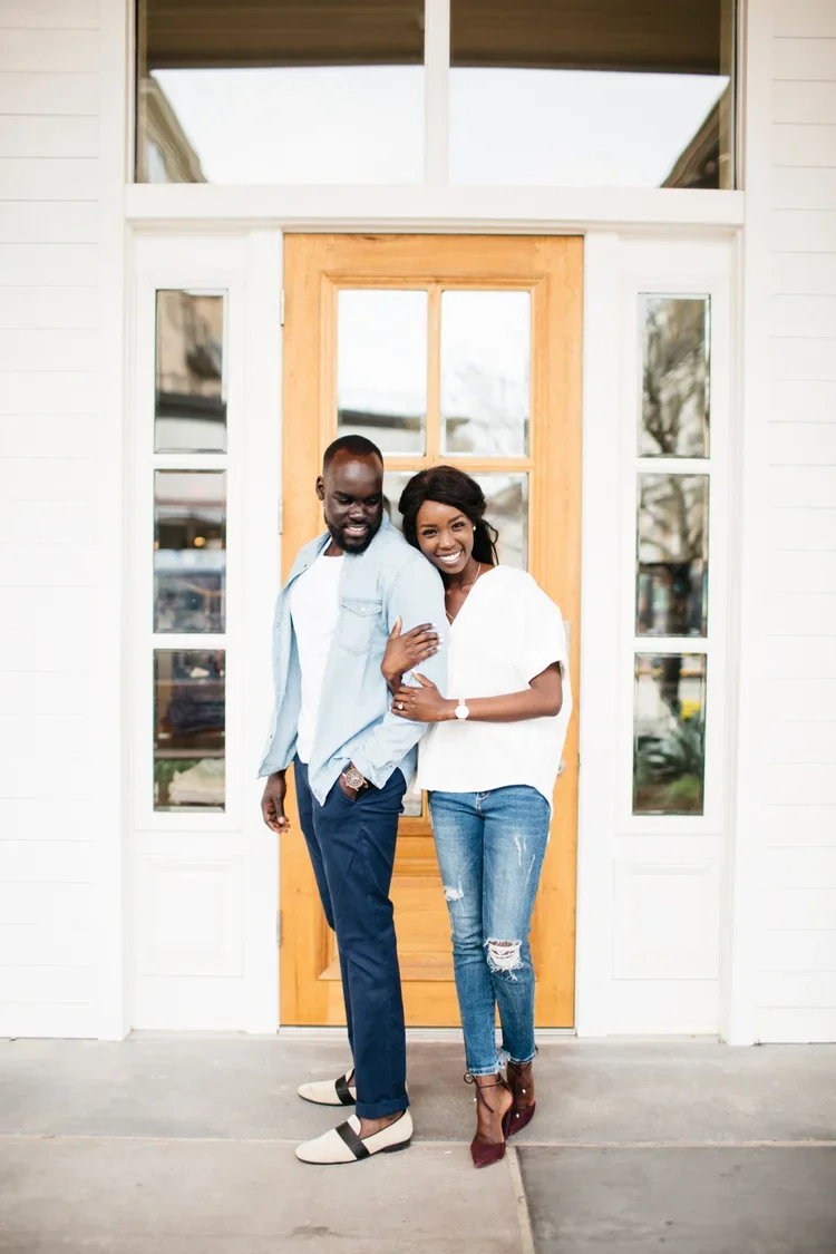A smiling woman and man standing arm in arm in front of a wooden door, outside a white house.