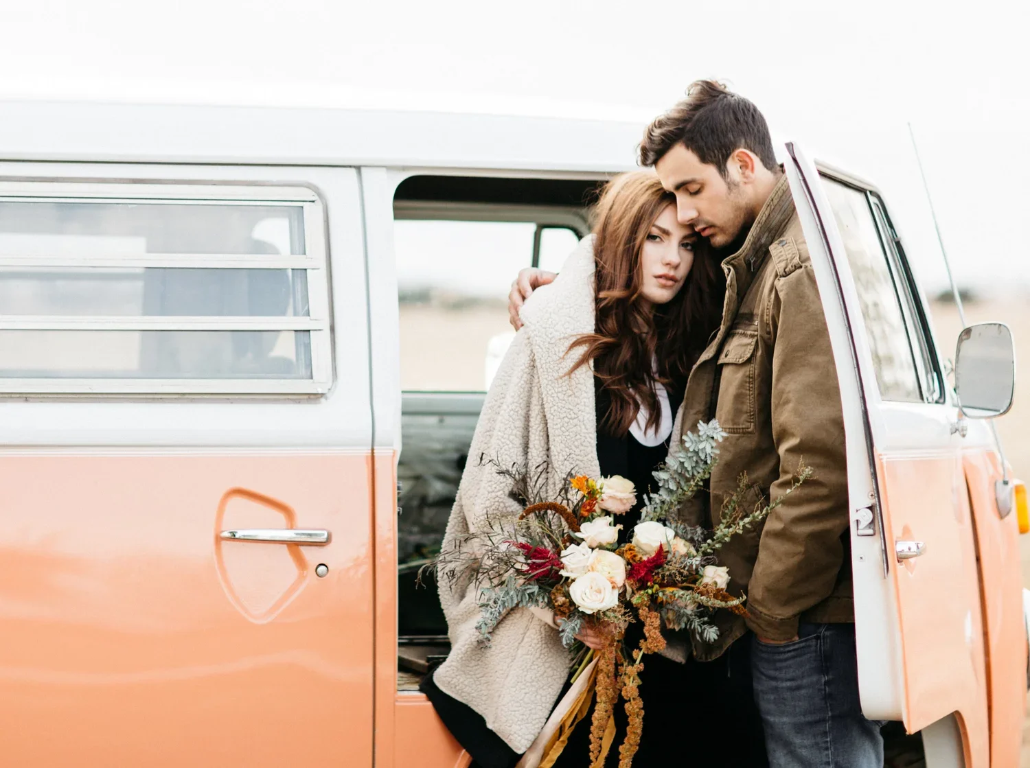 A young woman and a young man sharing an intimate moment beside a vintage orange and white van. The woman holds a bouquet of flowers, and they are adorned with a blanket and casual clothing, set in an outdoor, open field.