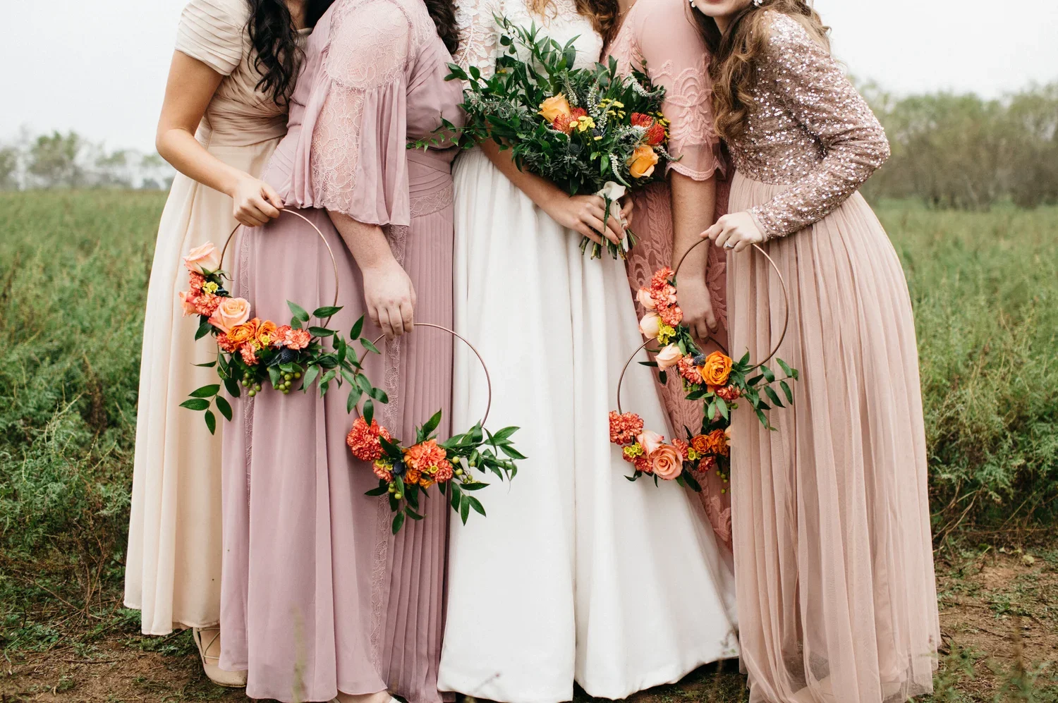 Four women in pastel-colored dresses standing outdoors, holding bouquets of flowers and floral crowns in a grassy field during daytime.