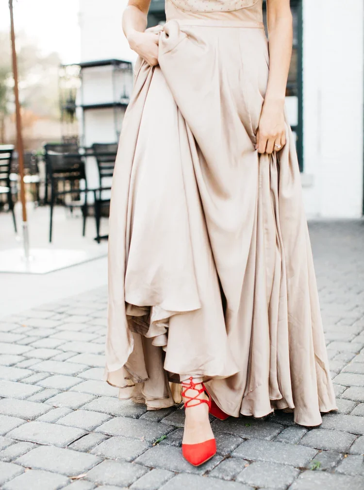 A woman wearing a beige gown and red heels standing on a cobblestone street.