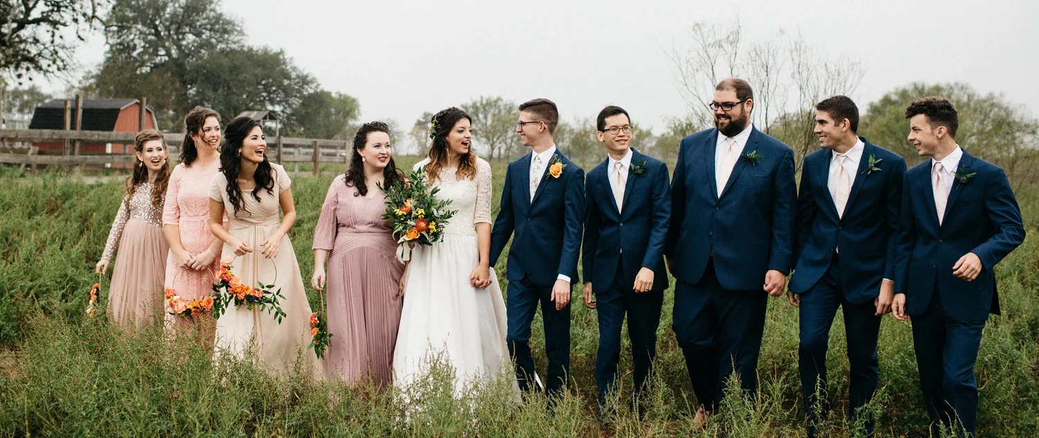 Group of people in formal attire walking outdoors on grassy field, with trees and a fence in the background, during a wedding celebration.
