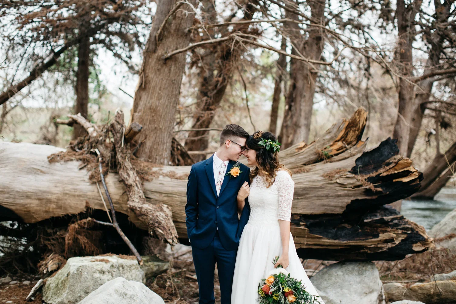 A bride and groom stand close together outdoors, touching foreheads and smiling, with a forest background and fallen logs.