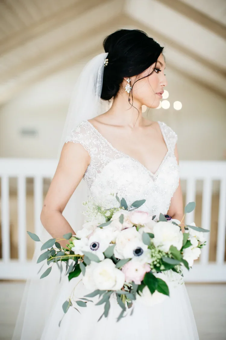 A bride in a white wedding dress holding a bouquet of flowers, standing indoors with a blurred background.