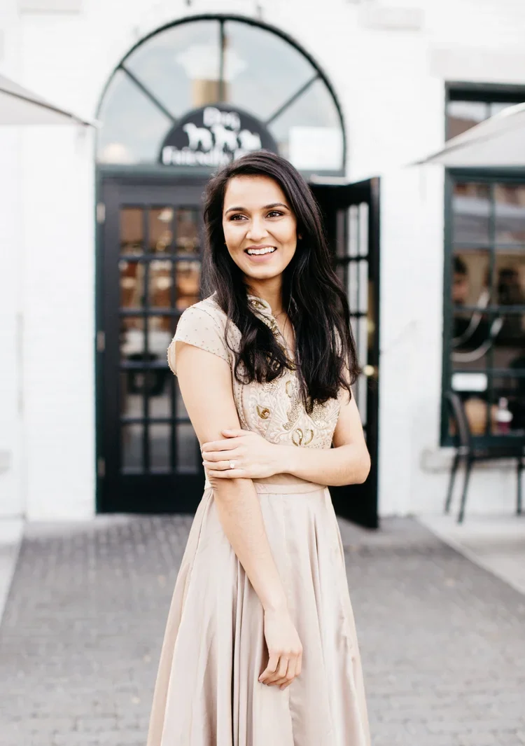 A young woman with long dark hair smiling, standing outside a building with a glass door and arched window.