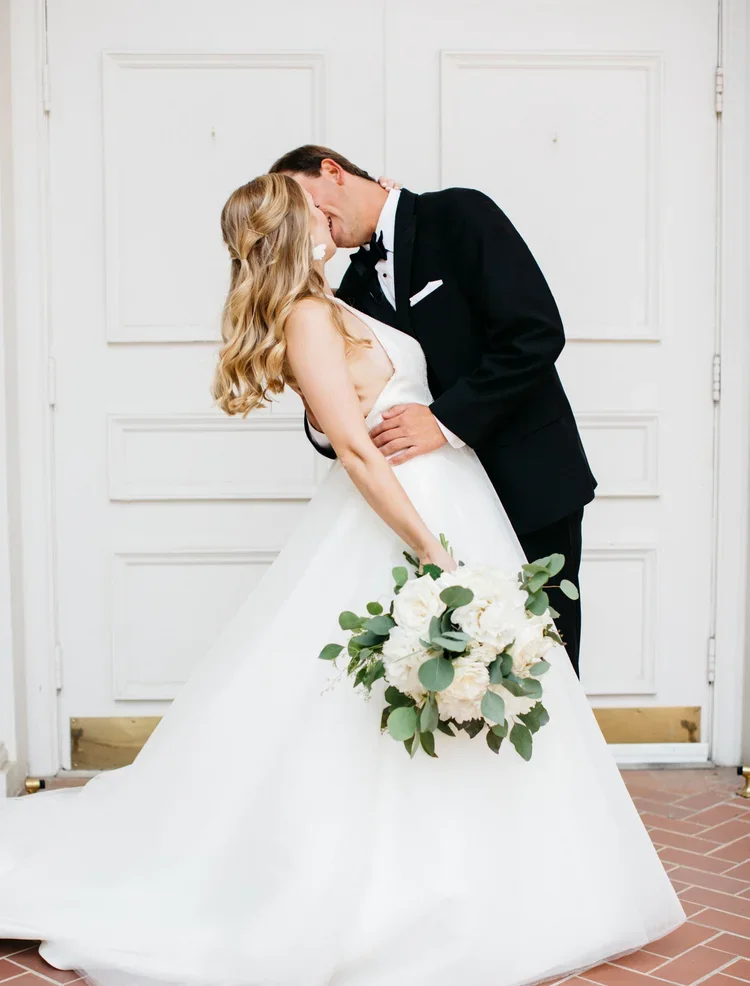 A couple dressed in wedding attire sharing a kiss indoors. The bride has long blonde hair and is holding a large bouquet of white flowers and greenery. The groom is wearing a black tuxedo and is embracing the bride. They are standing in front of a wh