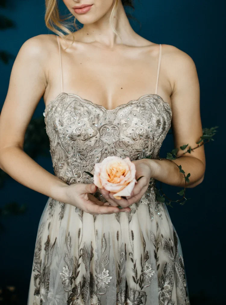 A woman in an ornate, strapless dress holding a pink rose with greenery, set against a dark background.