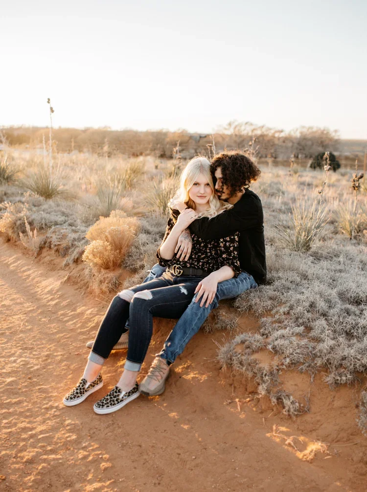A young couple sitting on a dirt path in a desert landscape with shrubs and cacti during sunset.