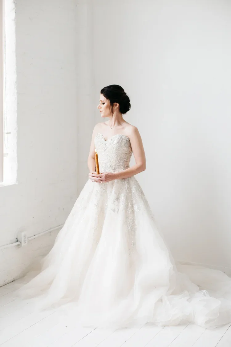 A woman in a white wedding gown with lace details, holding a bouquet, standing in a minimalistic room with white walls and floors.