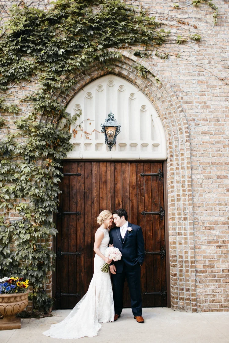 Bride and groom standing close together in front of a large wooden door with brick and ivy surroundings.