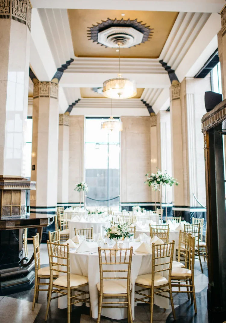 Elegant banquet hall with tall windows, high ceilings, and ornate columns. Round tables with white tablecloths and gold Chiavari chairs are decorated with floral centerpieces.