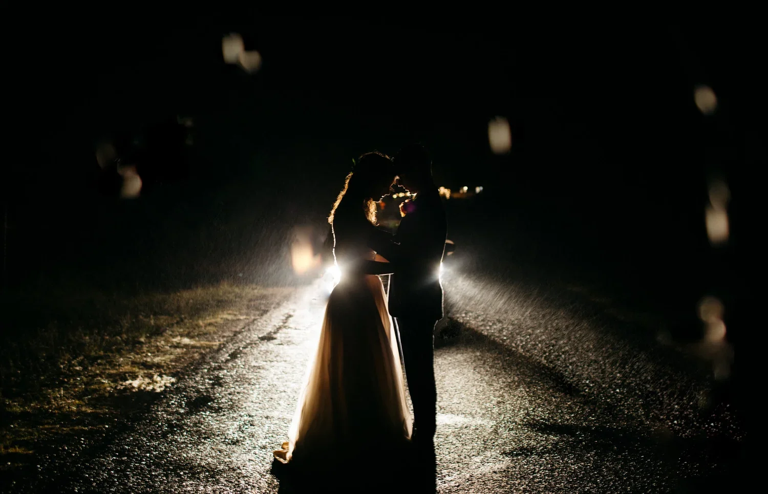 Silhouette of a couple kissing on a wet road at night, backlit by a bright light, creating a romantic scene.