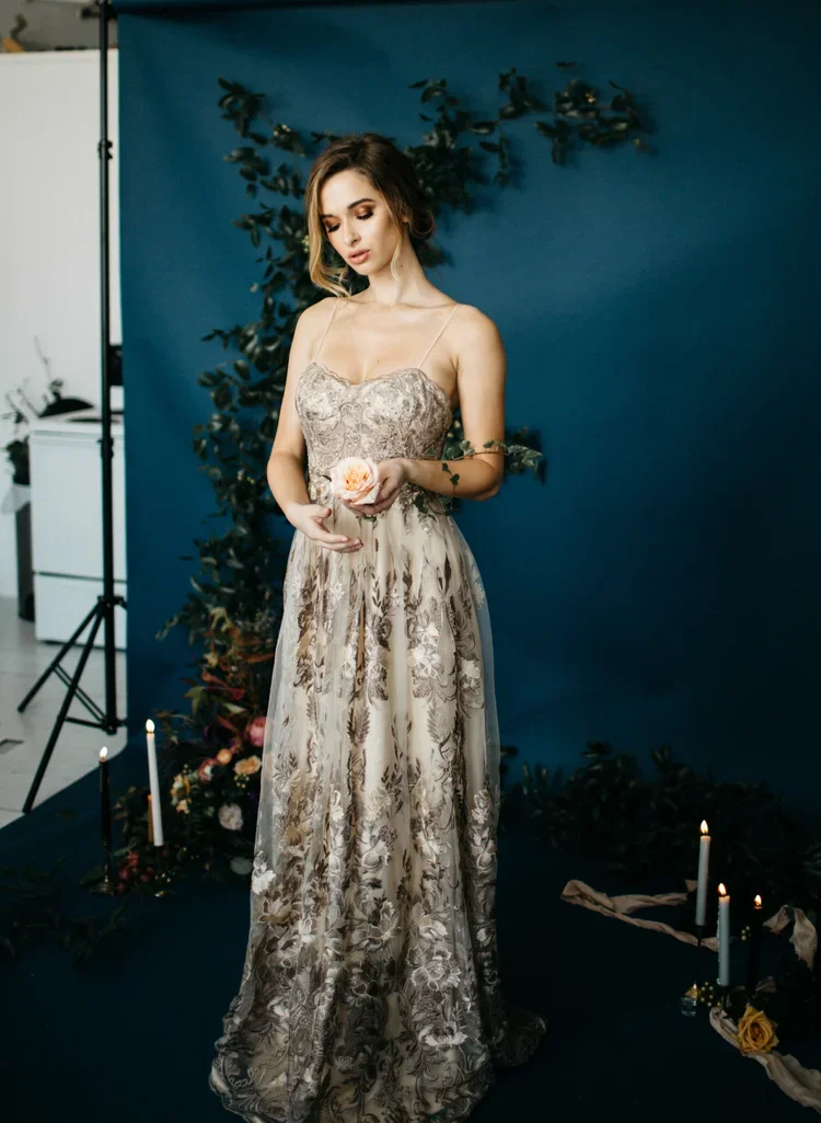 A young woman in a detailed, elegant floral dress standing against a dark blue backdrop, holding a flower, with candles and foliage on the floor.