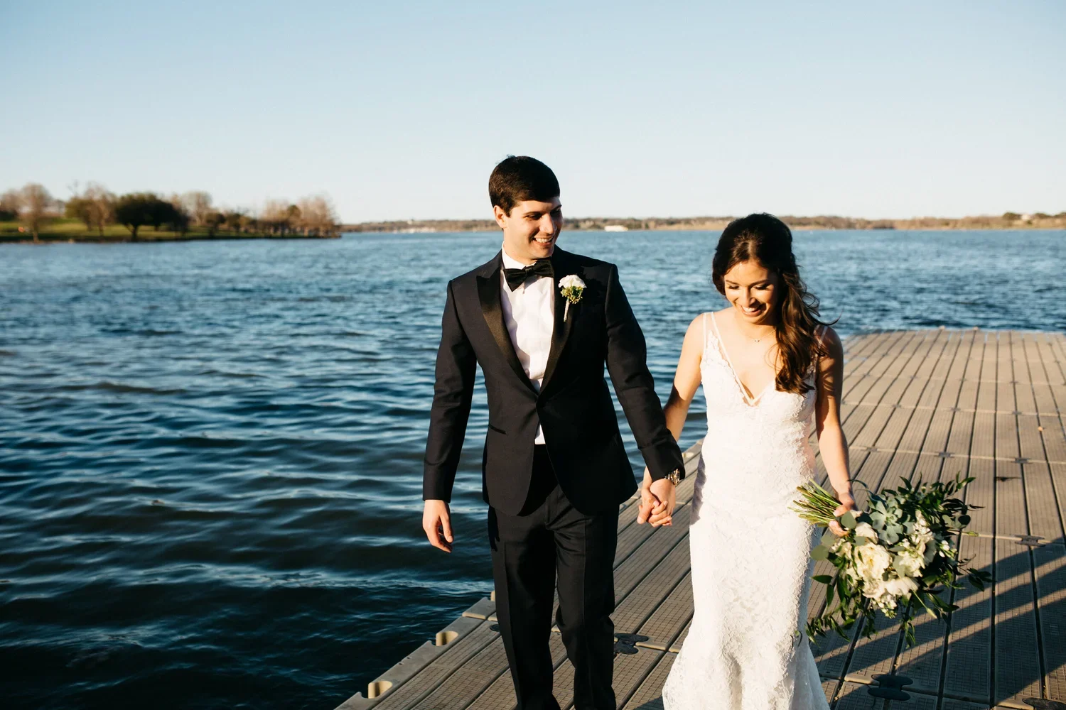 A newlywed couple walking on a wooden dock by a lake during sunset, holding hands and smiling. The groom is in a black tuxedo with a white shirt and bow tie, and the bride is in a white lace wedding dress holding a bouquet of flowers.