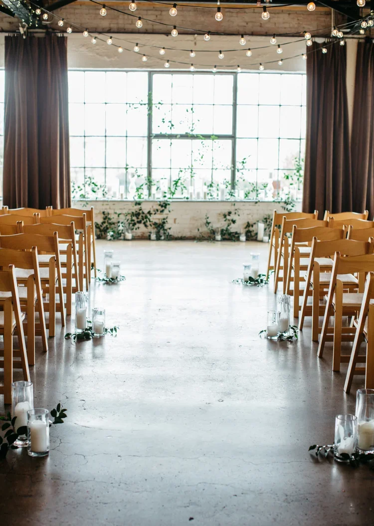 Empty wedding or event ceremony space with wooden chairs arranged on either side of an aisle decorated with candles and greenery, large industrial-style window at the back, string lights hanging from the ceiling, and curtains on the sides.