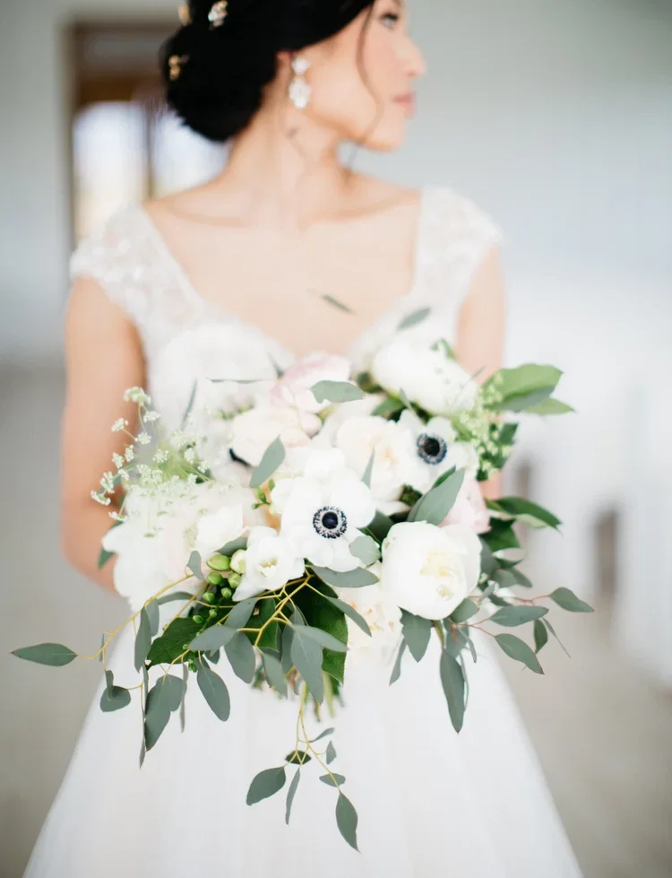 Bride holding a bouquet of white flowers and greenery, wearing a lace wedding dress and earrings, with blurred background.