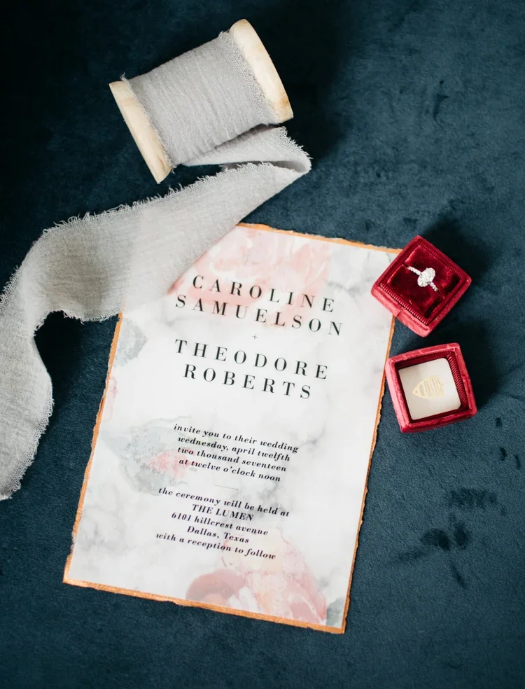 Wedding invitation with pink watercolor background, a spool of beige fabric, a red velvet ring box with a diamond ring, and a small red square box with a gold shell design.