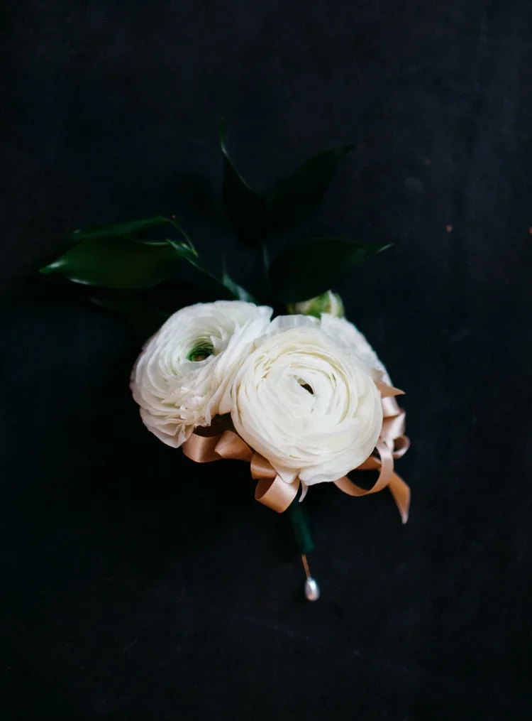 A boutonniere with white ranunculus flowers, dark green leaves, and a light brown ribbon against a black background.