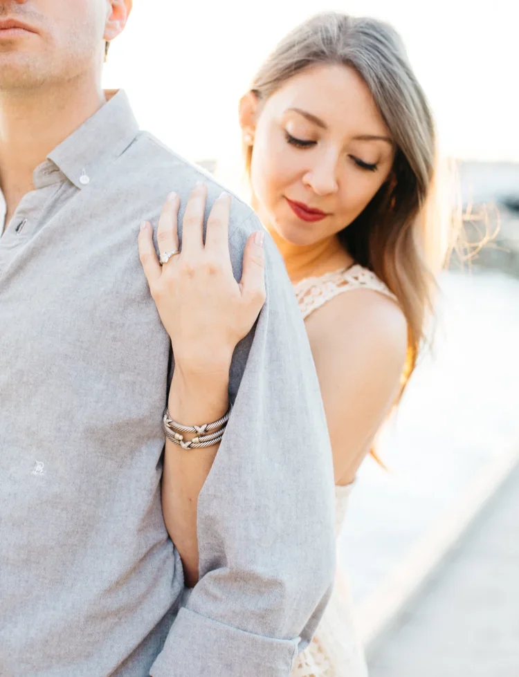 A woman gently rests her hand with an engagement ring on the shoulder of a man in a light gray shirt, standing outdoors during sunset.