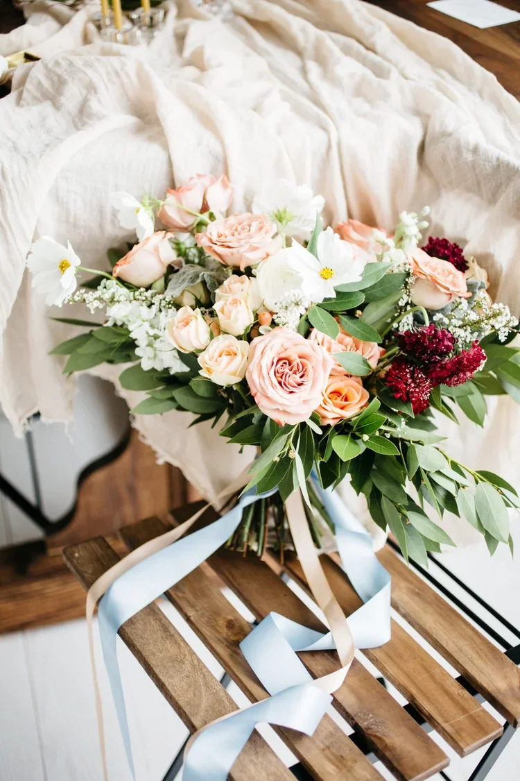 A bouquet of peach, white, and dark red flowers with green leaves and satin ribbons resting on a wooden bench.