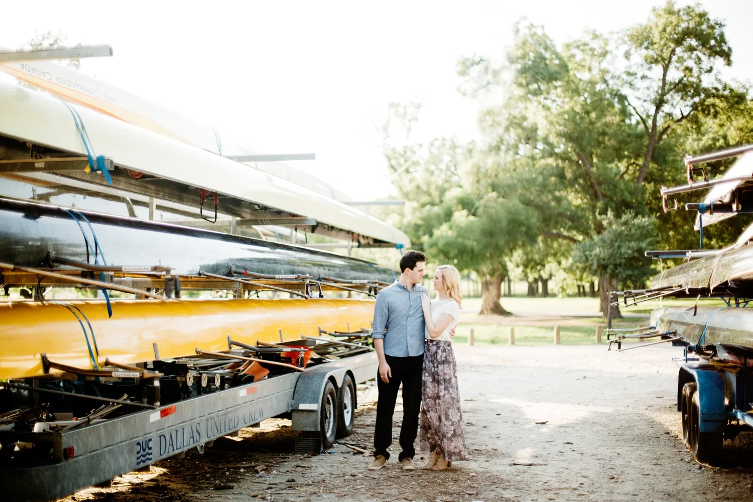 A young couple standing close together near boat trailers and canoes in a park, with trees and greenery in the background, during daylight.
