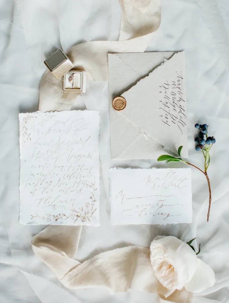 Elegant handwritten wedding invitations on textured paper, surrounded by a cream ribbon, a small ring box, a copper wax seal, a sprig of blueberries, a white rose, and soft fabric, all set on a white background.